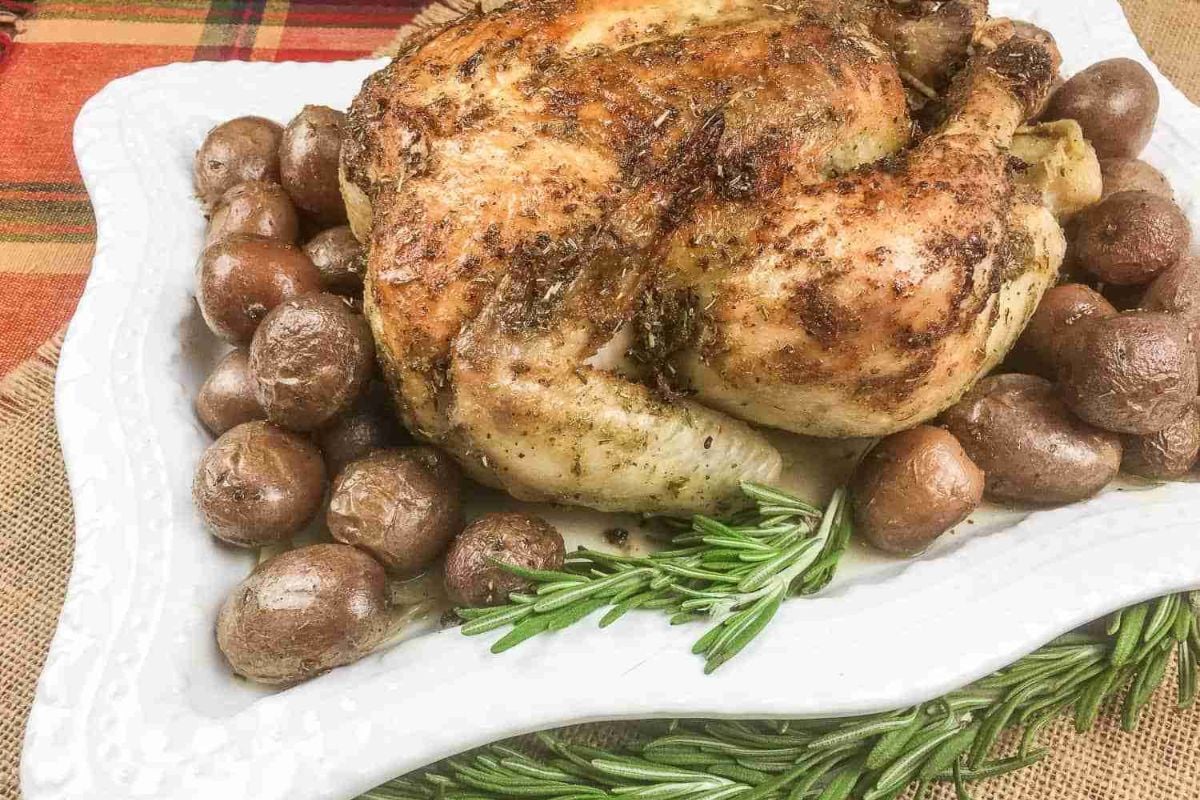 Roasted chicken with baby potatoes on a white plate, garnished with rosemary sprigs, placed on a patterned tablecloth.