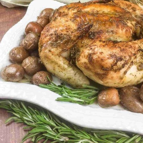 Roasted chicken with crispy skin is served on a white platter, surrounded by roasted potatoes and garnished with fresh rosemary sprigs.