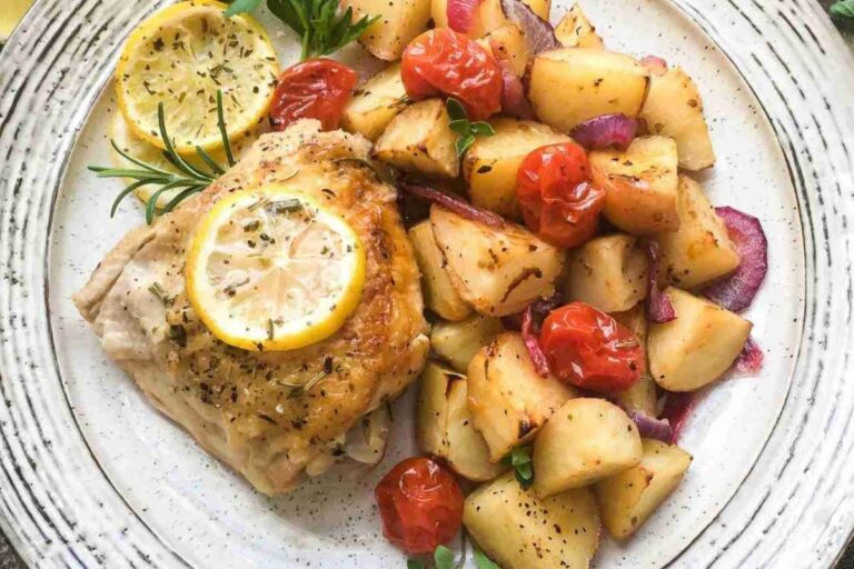 Plate with roasted chicken topped with lemon slices, served alongside roasted potatoes, cherry tomatoes, and red onions, garnished with herbs.