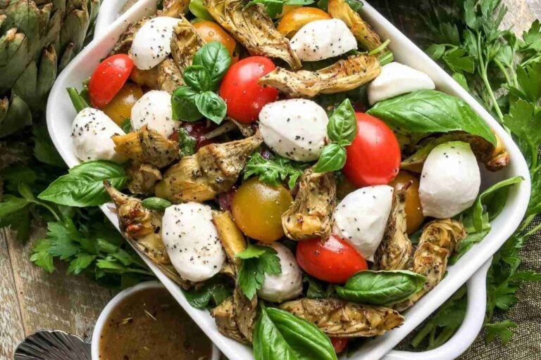 A dish of salad with cherry tomatoes, mozzarella balls, grilled artichoke hearts, and fresh basil leaves, garnished with black pepper.