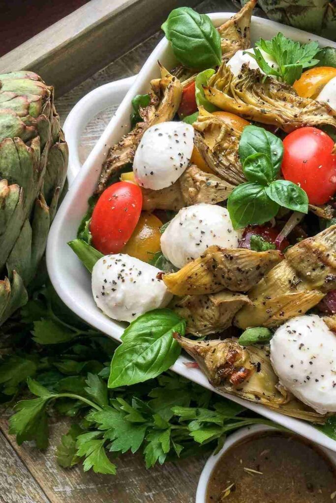 A salad with artichoke hearts, cherry tomatoes, mozzarella balls, and fresh basil leaves in a white dish, set on a wooden surface with herbs and an artichoke nearby.