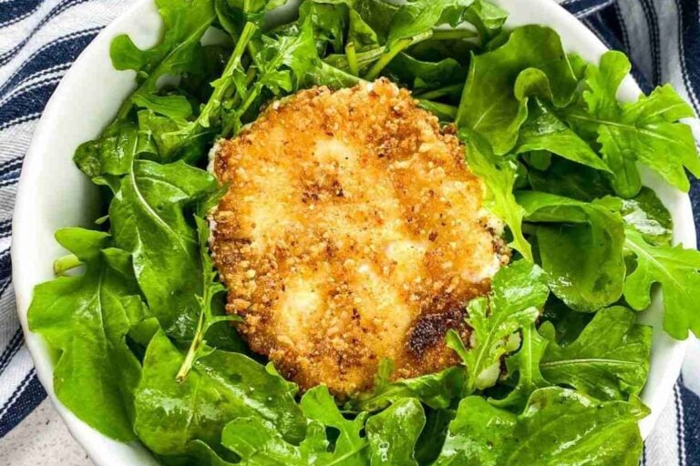 A breaded and fried cheese round sits on a bed of fresh arugula leaves in a white bowl, with a striped cloth partially visible underneath.