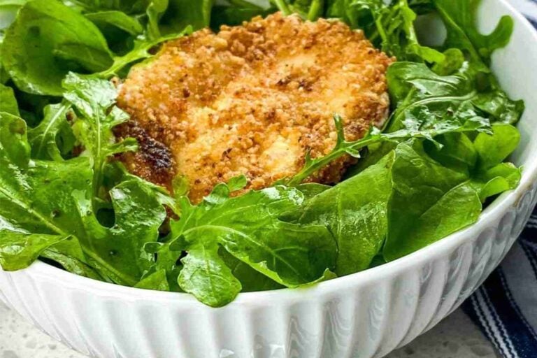 A white bowl filled with fresh green arugula leaves topped with a round, breaded and baked piece of cheese.