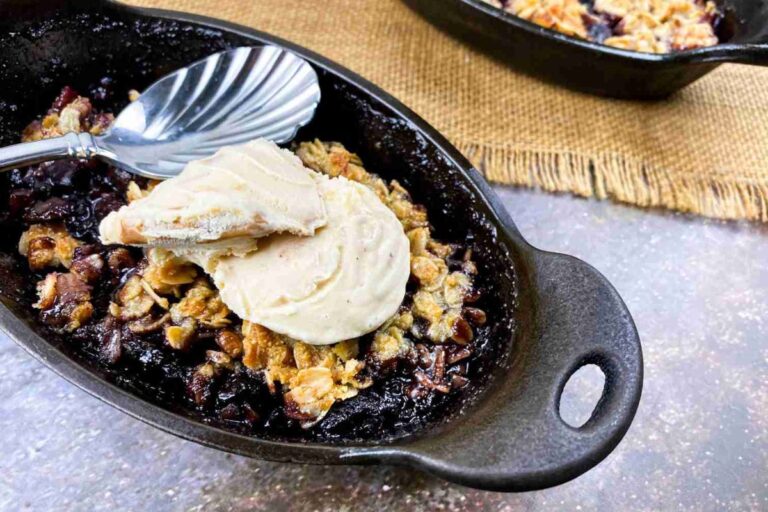 A small black dish holds a berry crumble topped with two scoops of vanilla ice cream and a spoon, placed on a textured surface.