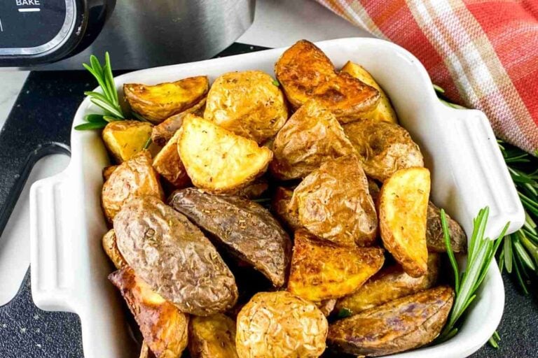 A white dish filled with roasted potato wedges, garnished with fresh rosemary, sits beside a plaid kitchen towel and a stainless steel appliance.