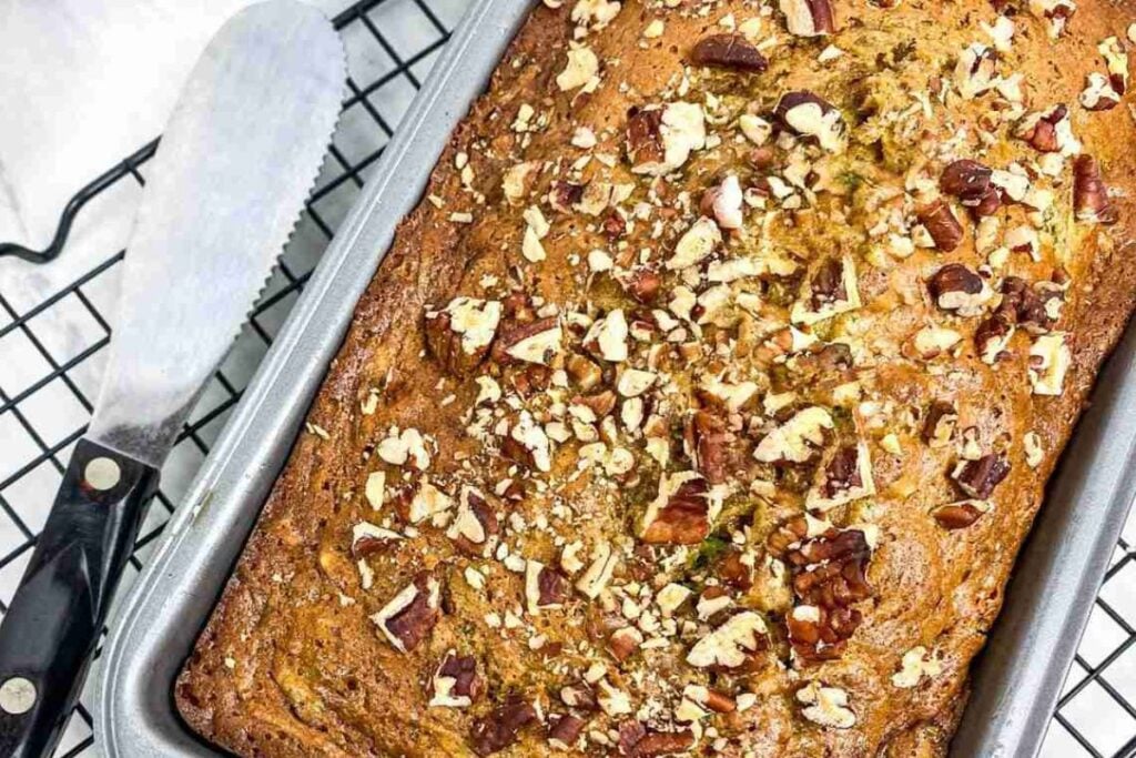 A loaf of banana bread topped with chopped pecans in a baking tin on a cooling rack, beside a serrated knife.