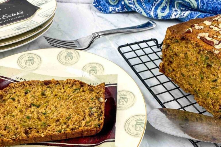 A sliced loaf of zucchini bread on a cooling rack with a piece served on a decorative plate beside a fork, over a marble countertop.