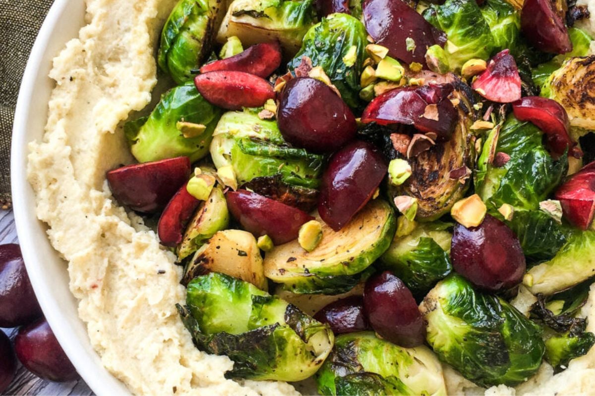 A bowl of mashed cauliflower topped with roasted Brussels sprouts, cherries, and chopped pistachios.
