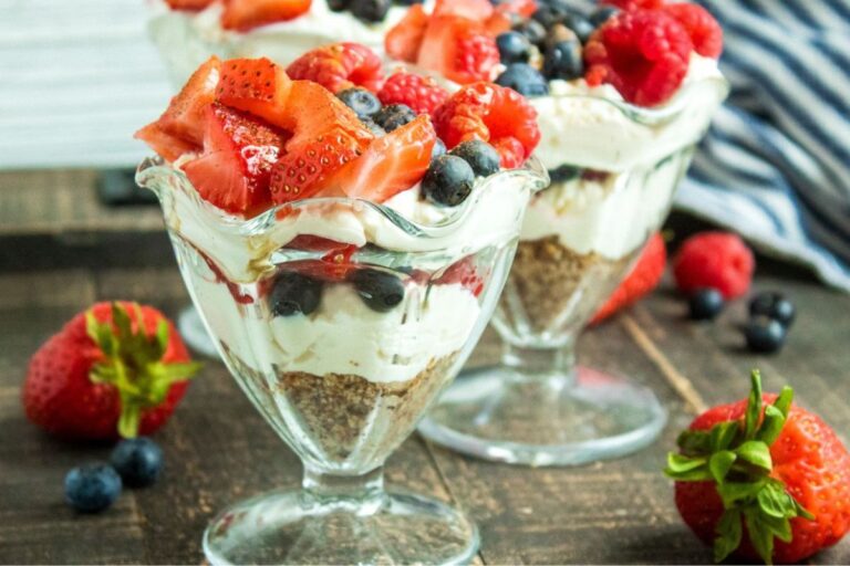 Two glass cups filled with layered dessert of cream, crushed biscuits, and mixed berries, including strawberries, blueberries, and raspberries, on a wooden table.