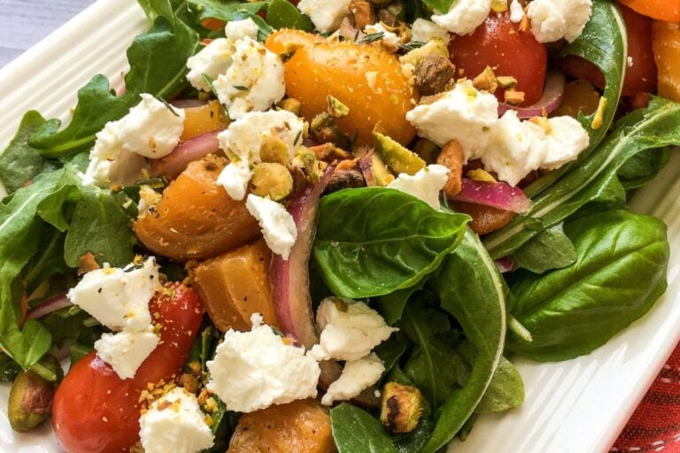 A fresh salad with arugula, spinach, roasted golden beets, cherry tomatoes, sliced red onions, goat cheese crumbles, and chopped pistachios on a rectangular white plate.