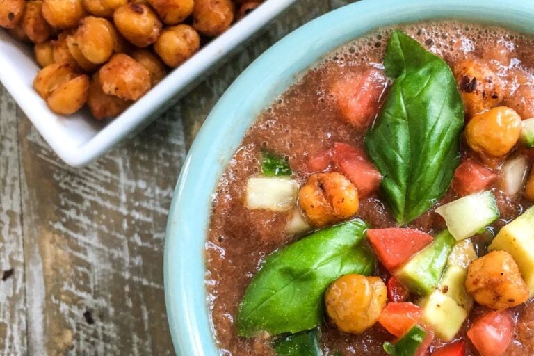 A bowl of vegetable soup with chickpeas, tomatoes, cucumbers, and basil leaves, next to a small dish of roasted chickpeas on a wooden table.