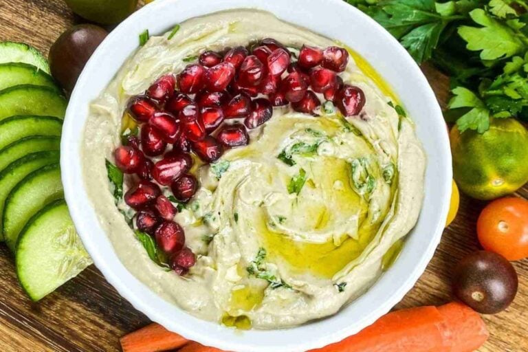 A bowl of hummus garnished with pomegranate seeds and olive oil, surrounded by sliced cucumber, carrots, tomatoes, fresh herbs, and a side of creamy baba ganoush.