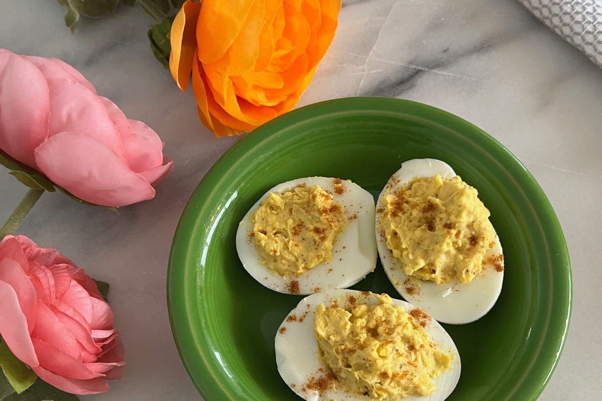 Three of grandma's deviled eggs rest on a green plate, garnished with paprika. Nearby, pink and orange flowers adorn the marble surface.