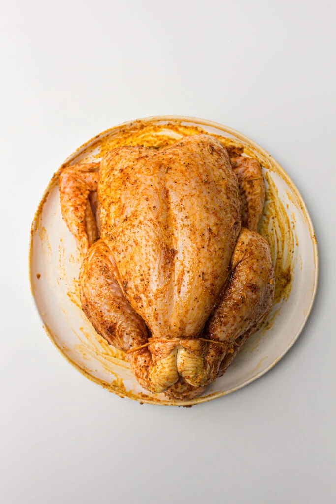 A raw, seasoned whole chicken on a white plate with a beige rim, viewed from above, ready for cooking in an air fryer.