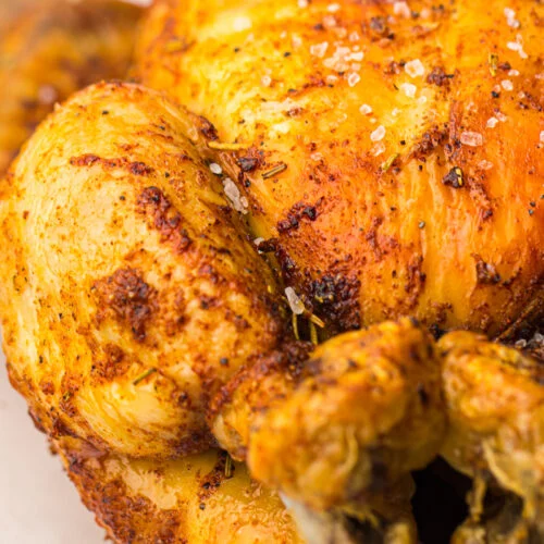 Close-up of a whole chicken roasted in an air fryer with crispy, golden-brown skin, seasoned with herbs and spices on a wooden board.