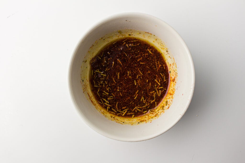 A white bowl containing a reddish-brown sauce with visible herbs and spices, accompanied by a whole chicken in an air fryer, on a bright white background.