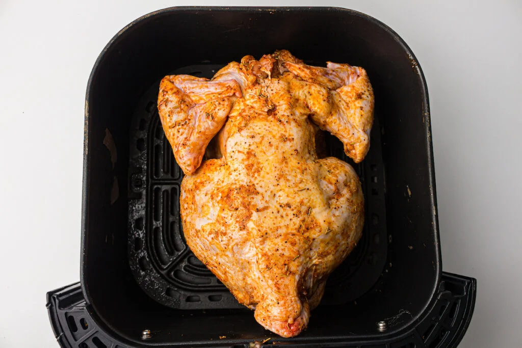 A whole chicken in an air fryer basket, seasoned and ready to be cooked.