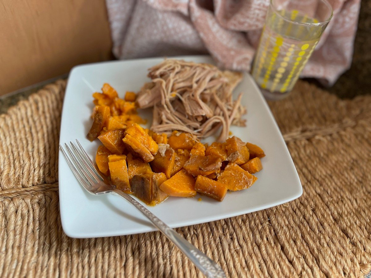 A plate of sliced roast pork loin and cubed sweet potatoes, with a fork on the plate and a glass of water on a woven placemat.