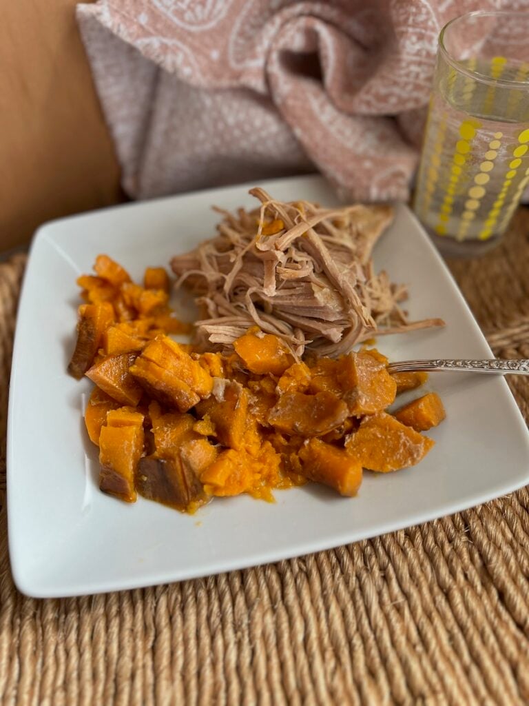 A plate of pulled pork and diced sweet potatoes, with a patterned napkin and a glass of water beside it on a textured mat.