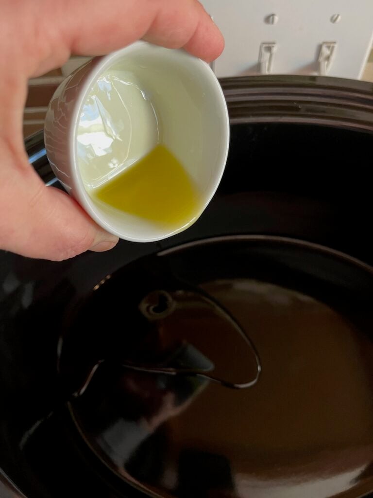 A person pouring olive oil from a small cup into a black cooking pot.