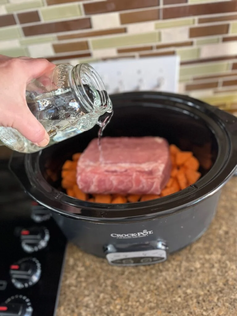 A person pours water from a jar into a slow cooker containing carrots and a pork roast.