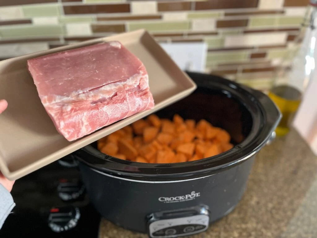 A person holding a raw pork roast above a slow cooker filled with carrots.