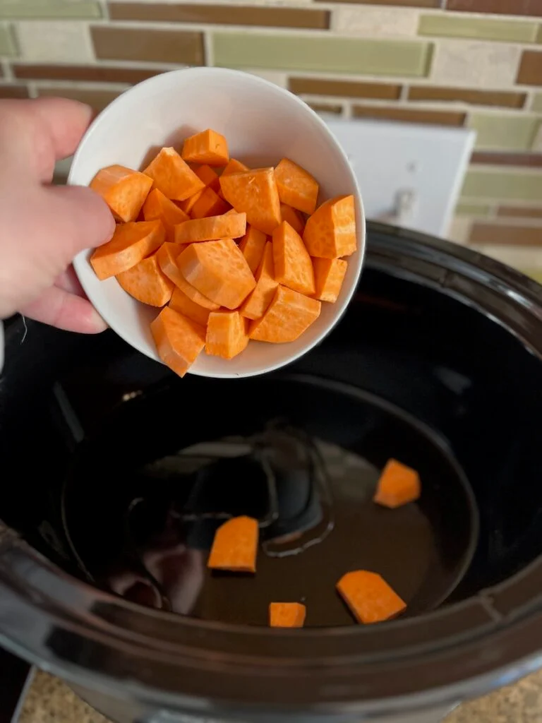 A person holding a white bowl filled with chopped sweet potatoes, about to pour them into a black slow cooker.