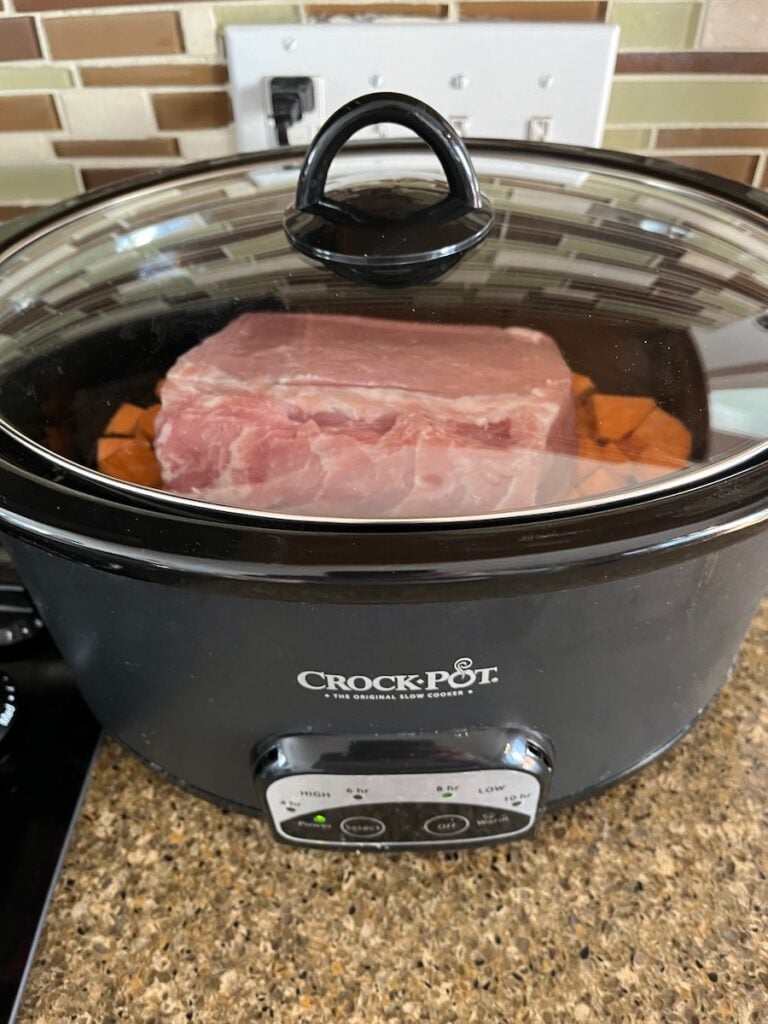 A large piece of raw pork sits inside a black crock-pot slow cooker on a kitchen counter, surrounded by chopped vegetables.