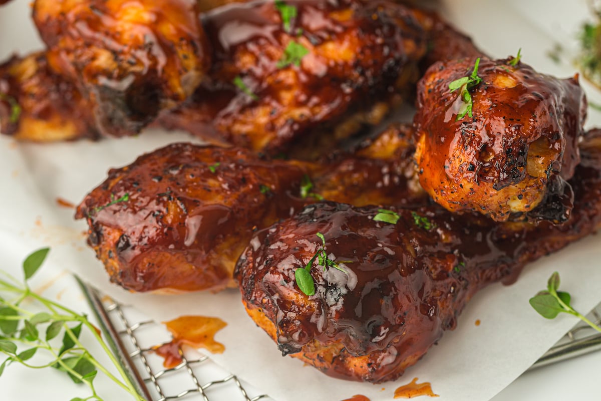 Close-up of barbecue chicken drumsticks in the air fryer, glazed with a glossy, dark red sauce, garnished with fresh green herbs, served on a piece of white parchment paper.