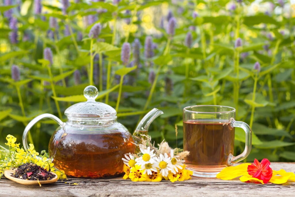Glass teapot and cup with herbal tea, surrounded by colorful wildflowers on a wooden table outdoors.