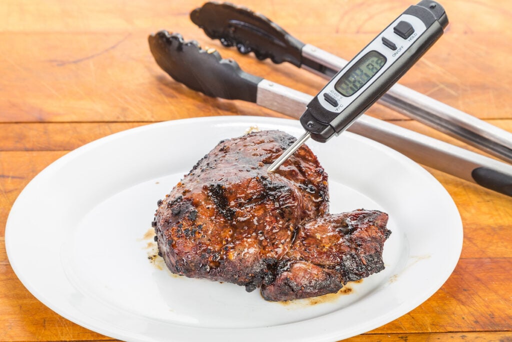 A cooked piece of seasoned meat on a white plate with a digital thermometer inserted into it, resting on a wooden surface with metal tongs placed behind the plate.
