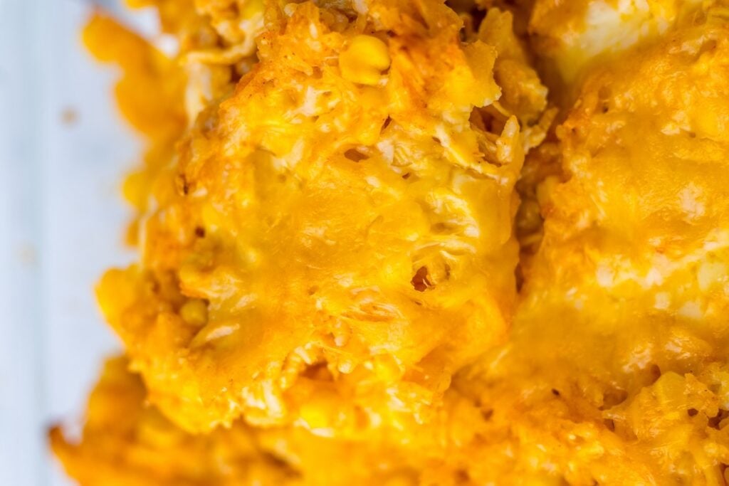 Close-up of a cheesy casserole with visible golden melted cheese, corn kernels, and chunky pieces.