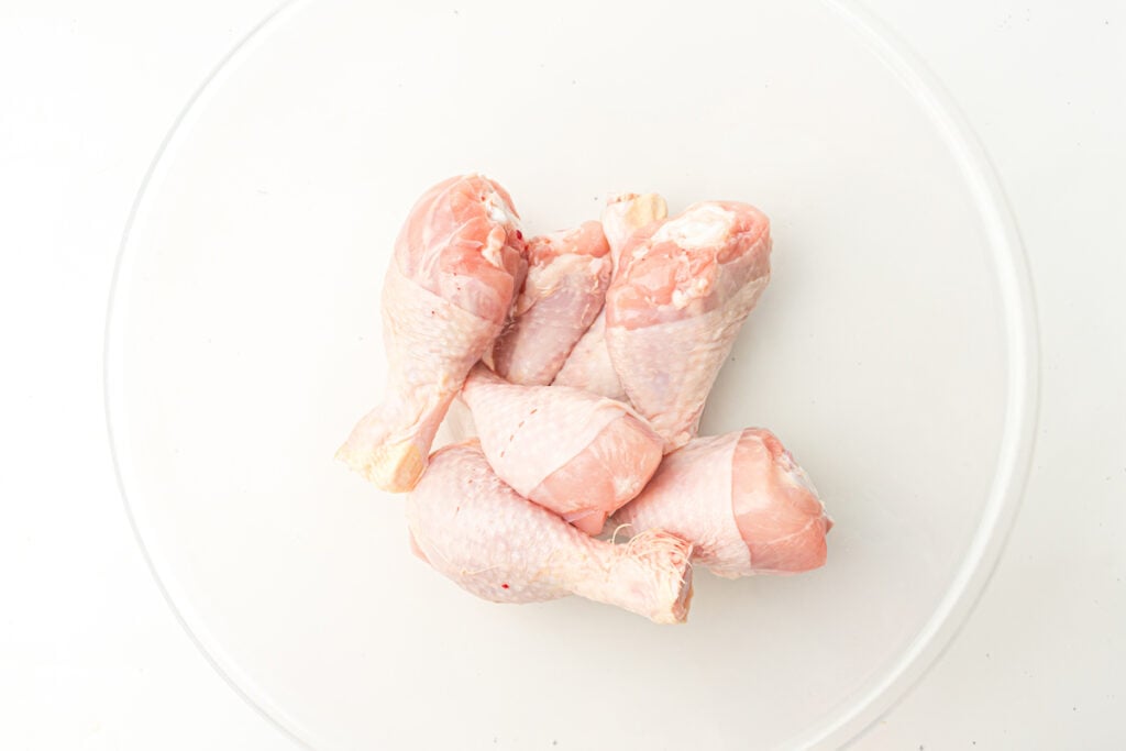 A clear bowl containing five raw chicken drumsticks on a white background.