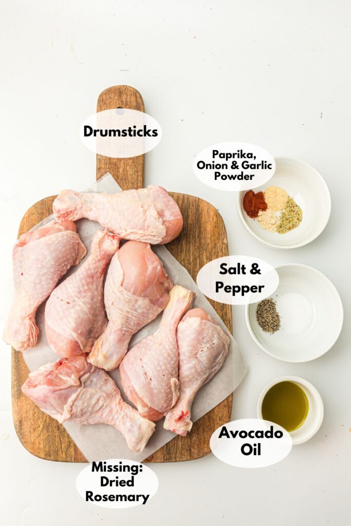 Raw chicken drumsticks on a wooden board with small bowls of paprika, onion powder, garlic powder, salt, pepper, and avocado oil. Note indicates missing dried rosemary.