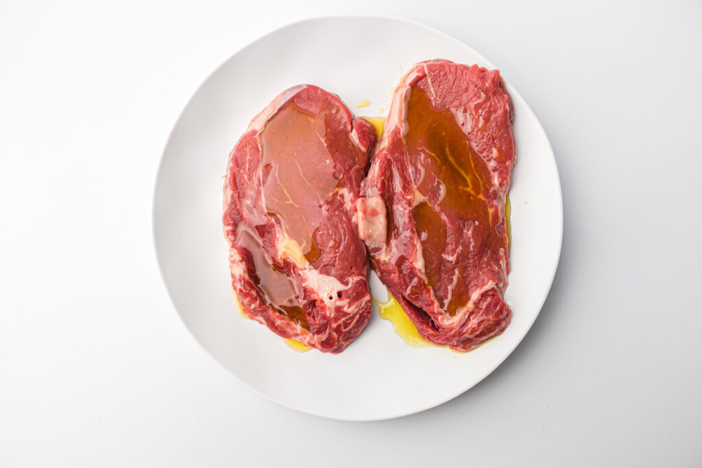 Two raw beef steaks on a white plate, drizzled with olive oil, placed on a white background.