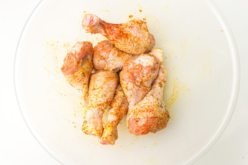 Raw chicken drumsticks seasoned with spices are placed in a clear glass bowl.