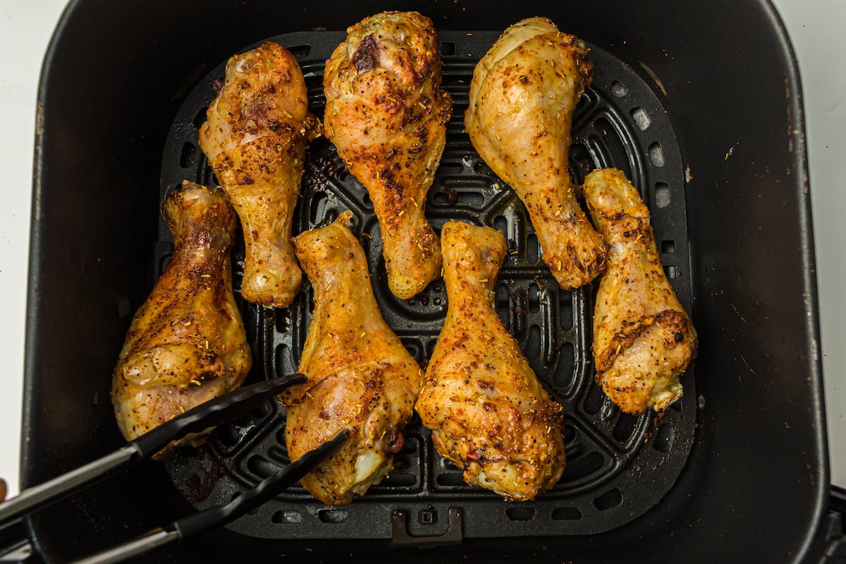Six seasoned chicken drumsticks are shown being cooked in an air fryer basket, with a pair of tongs holding one.