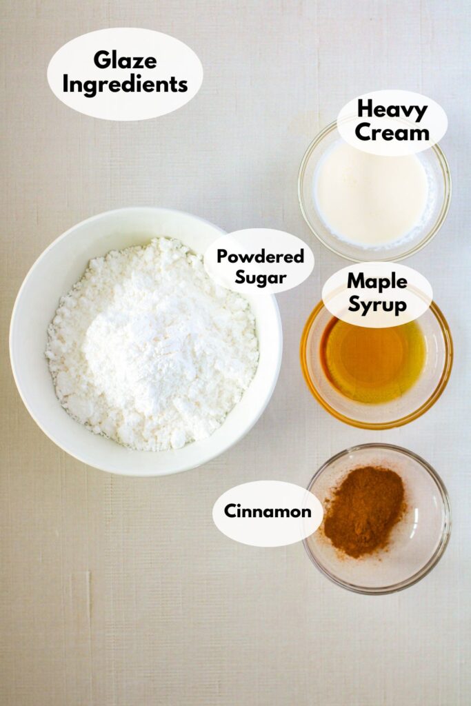 Ingredients for a glaze are arranged in bowls on a surface, including powdered sugar, heavy cream, maple syrup, and cinnamon. Each ingredient is labeled in the image.
