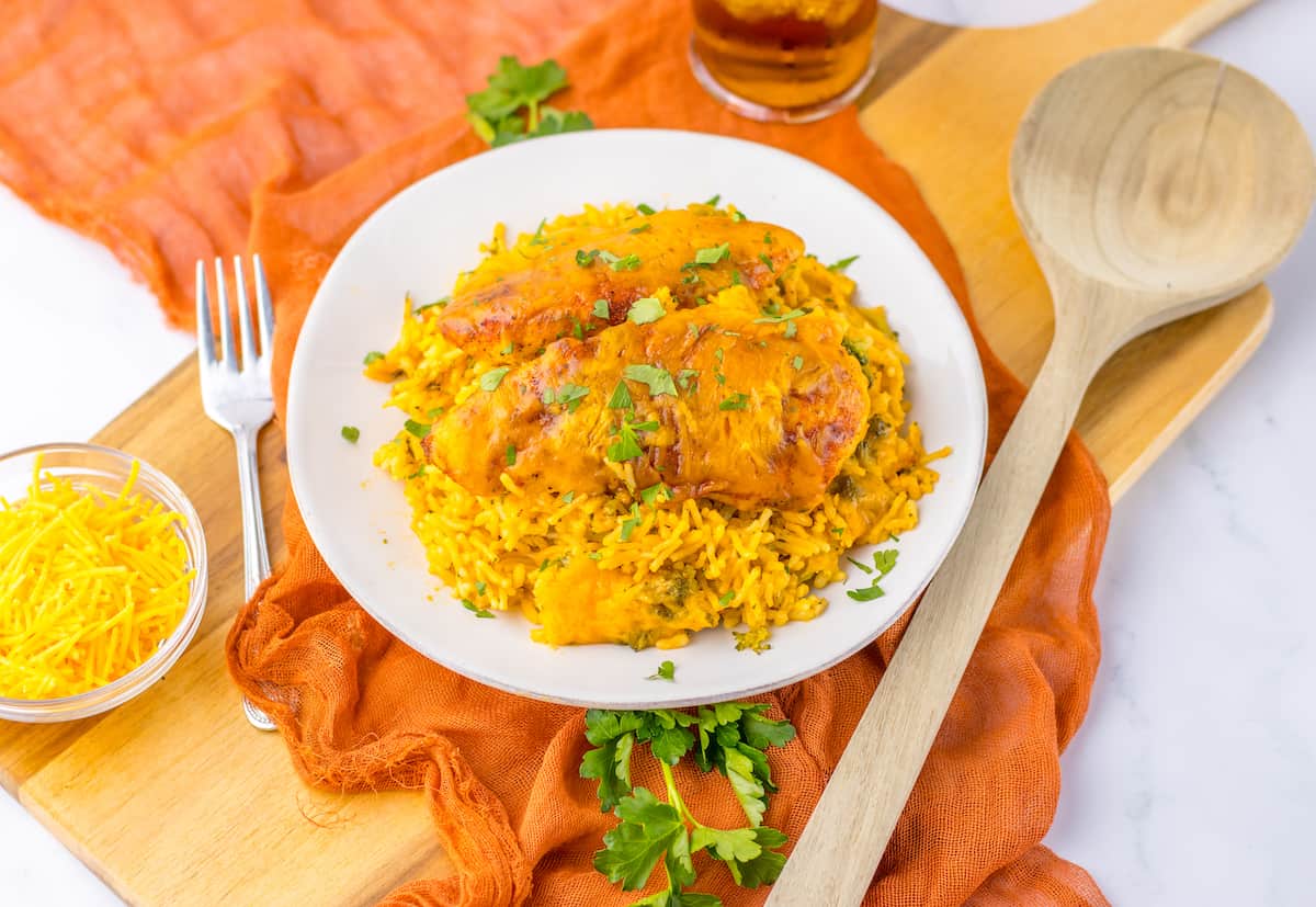 A plate of chicken with rice garnished with parsley on a white surface with an orange napkin, wooden utensils, and ingredients nearby.
