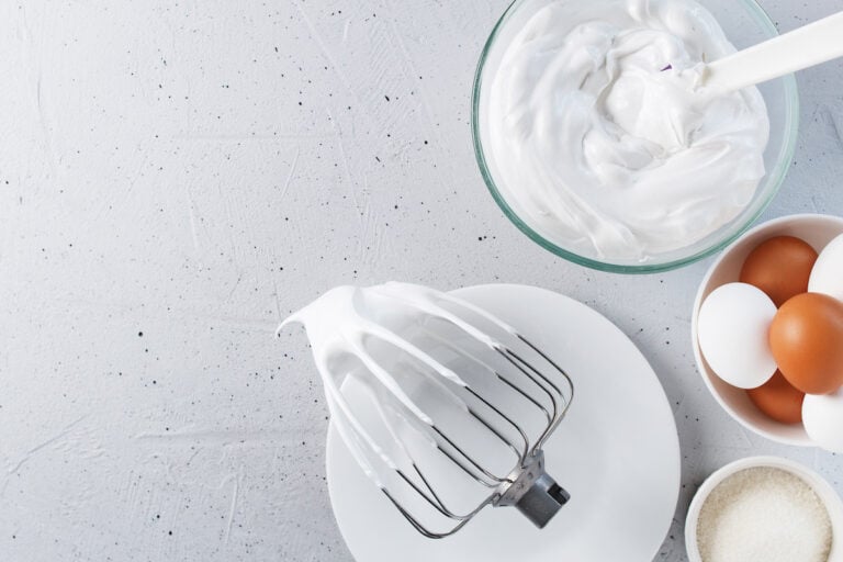A beater covered in white whipped cream rests on a plate next to a bowl of whipped cream, eggs in a bowl, and a small bowl of granulated sugar on a light textured surface, illustrating the first steps of how to make meringue.