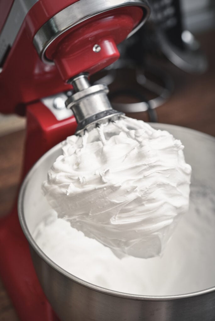 A red stand mixer with a whisk attachment holds a large amount of whipped cream in a metal mixing bowl, perfect for those learning how to make meringue.