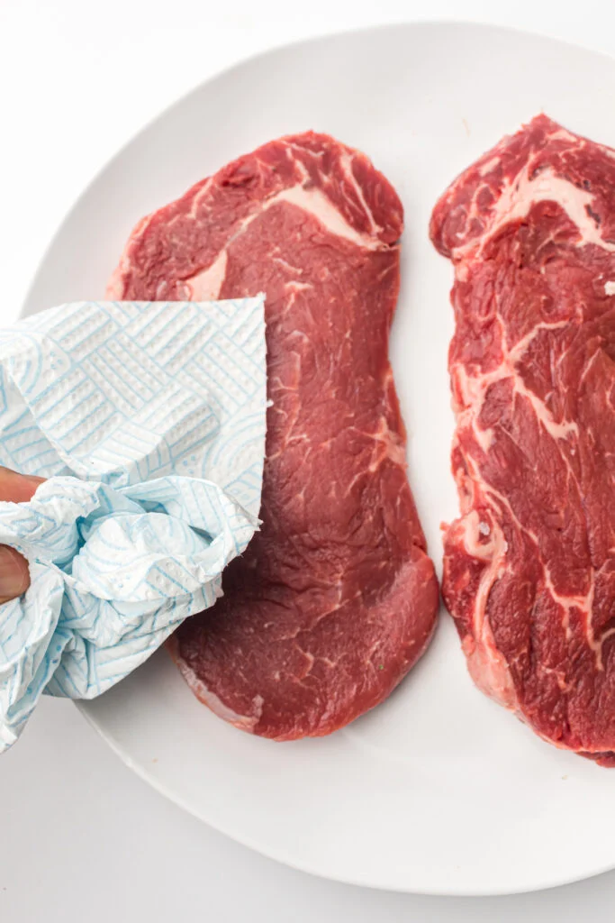 A hand uses a paper towel to pat dry two raw steaks on a white plate.