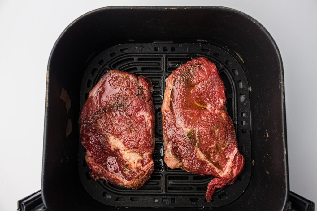 Two raw, seasoned steaks placed inside an air fryer basket, ready for cooking.