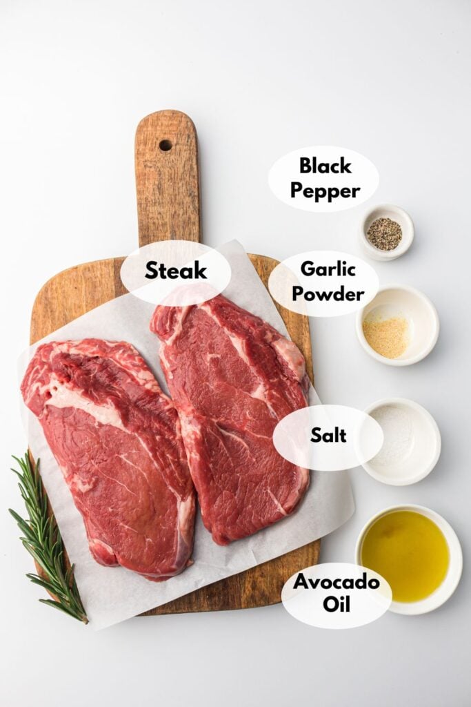A wooden board with two raw steaks, rosemary, and labeled ingredients: black pepper, garlic powder, salt, and avocado oil, displayed in small white bowls on the side.