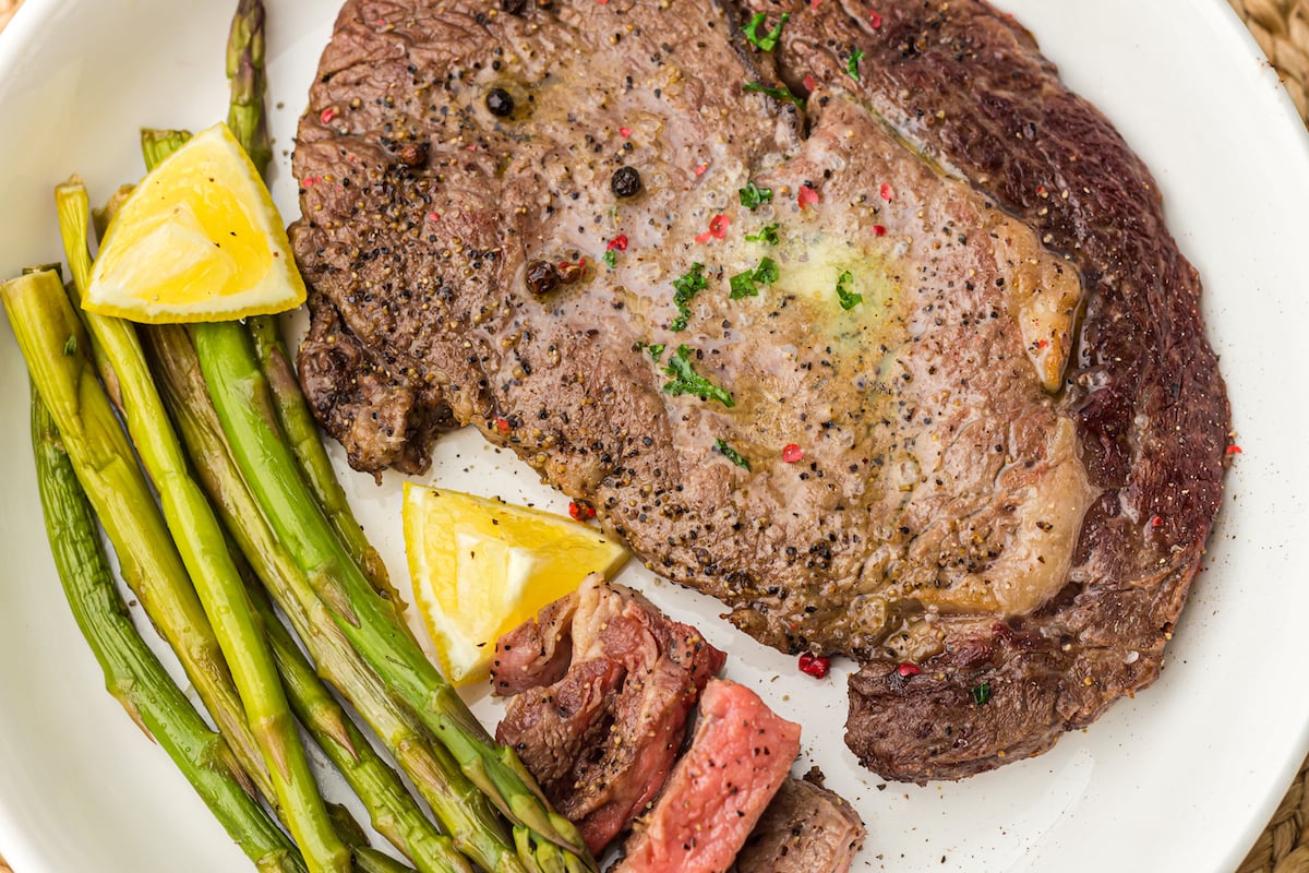 A plate with a grilled steak topped with herbs and black pepper, served with asparagus spears and lemon wedges.