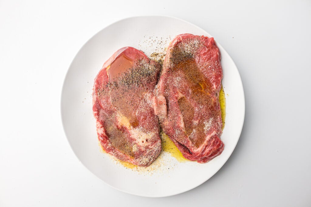 A white plate with two raw steaks seasoned with salt, pepper, and oil, resting on a white background.
