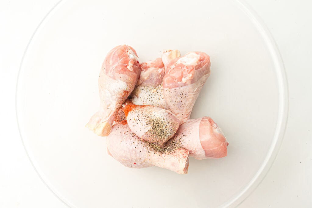Raw chicken drumsticks seasoned with salt, pepper, and paprika placed in a white bowl.