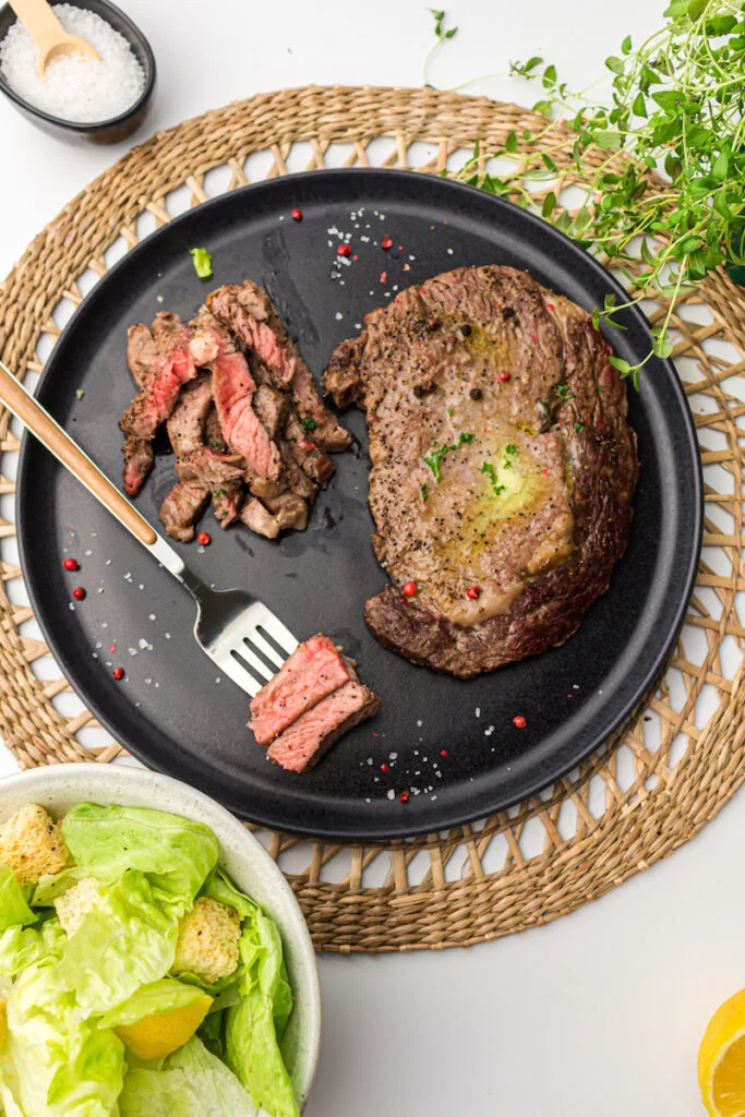 A cooked steak with a few cut pieces on a black plate, a fork, a side salad with croutons in a bowl, and a small bowl of salt on a woven placemat.