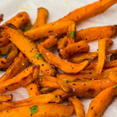 Close-up of a pile of seasoned sweet potato fries garnished with small green herbs, served on a white plate—perfect for celebrating National French Fry Day.