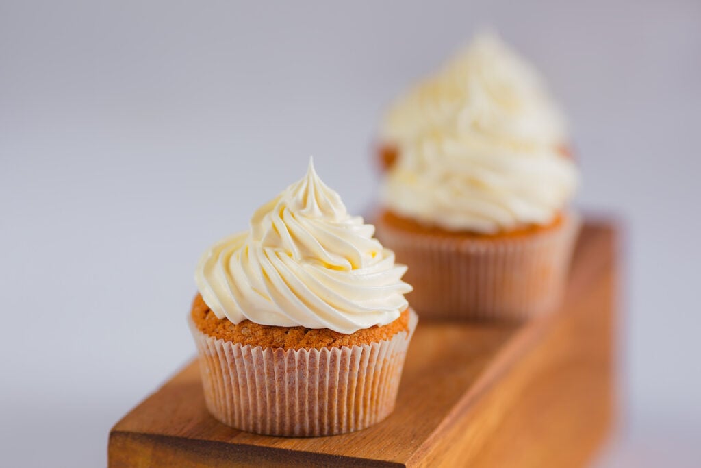 Two cupcakes with swirled white frosting, reminiscent of how to make meringue, sit on a wooden board against a plain background.
